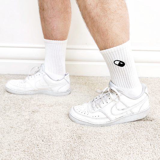 Side view of a model wearing the white socks paired with white trainers, showing the visible pill embroidery above the shoe.