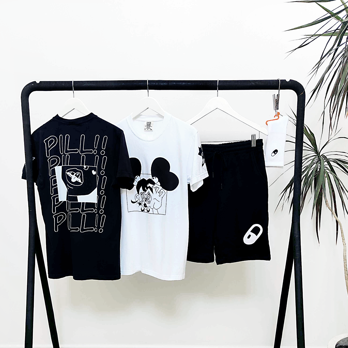 Collection 2 The Pill lifestyle mock up of the black and white t-shirts and shorts on a rack with a plant in the background.