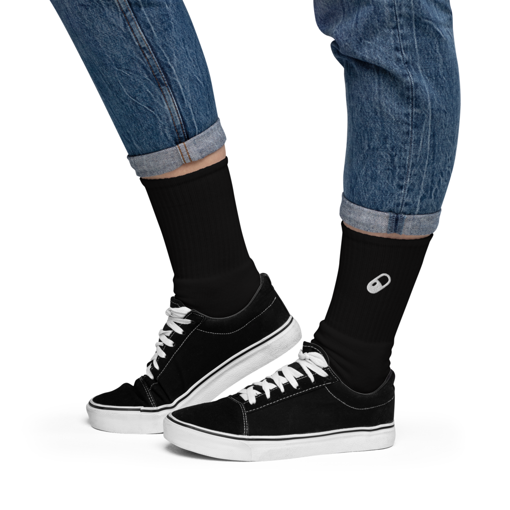 Side view of the model wearing black socks with black ankle canvas shoes, with the embroidered pill peeking out above the shoe.