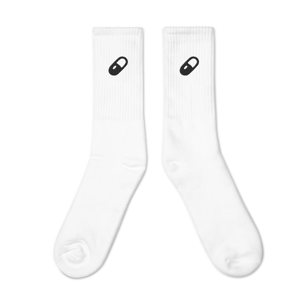 Flat lay of white crew socks with a thick black embroidered pill design on the side of each sock.
